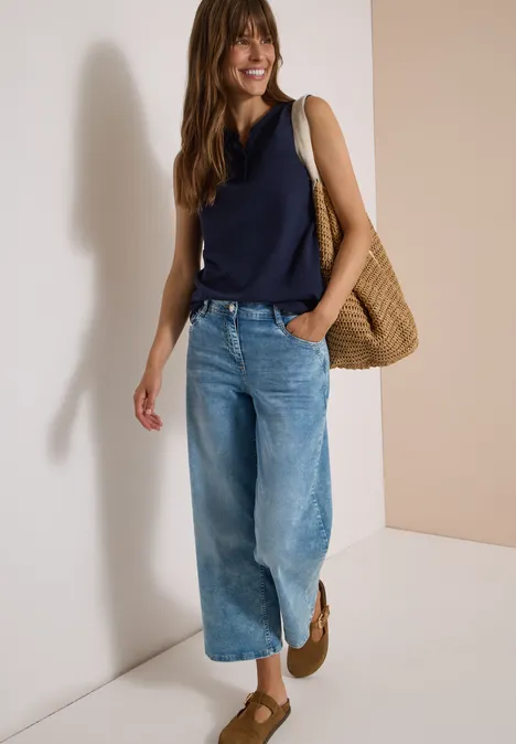 Frau steht an heller Wand, trägt dunkelblaues Top, weite Jeanshose, Basttasche und braune Schuhe.
