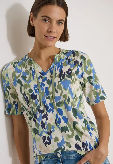 T-Shirt mit Split Neck und Print cashmere beige Frau trägt kurzärmliges Oberteil mit blau-grün-weißem abstraktem Muster.