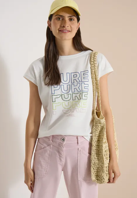 Frau mit weißem T-Shirt 'PURE', rosa Hose und gehäkelter Strandtasche.