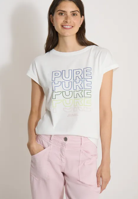 Junge Frau vor weißem Hintergrund trägt weißes T-Shirt mit „PURE“ Aufdruck in verschiedenen Farben und rosafarbene Hose.