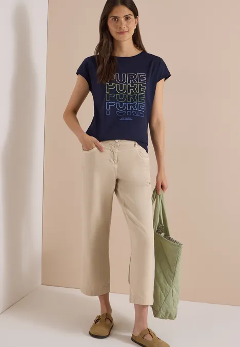 Frau in blauem Print-T-Shirt, beige Culotte, olivgrüner Tasche und beigen Slippern.