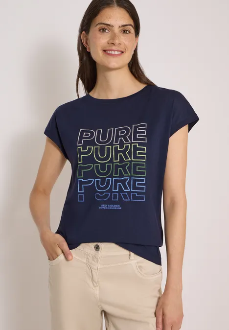 Frau in dunkelblauem T-Shirt mit 'PURE YUKE'-Schriftzug und beigen Hosen steht entspannt.