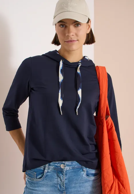 Eine Person trägt dunkelblauen Kapuzenpullover, Jeans, roten Stoffbeutel und Baseballcap vor hellem Hintergrund.