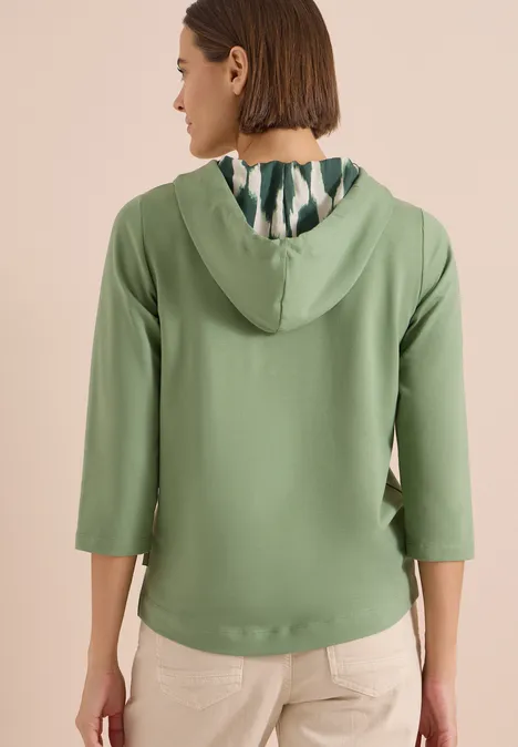 Frau von hinten in salbeigrünem Kapuzenpullover mit 3/4-Ärmeln und gemusterter Kapuzeninnenseite.