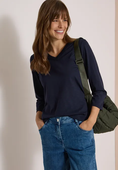 3/4-Arm Shirt in Unifarbe mit V-Neck urban dark blue Frau mit dunkelblauem Pullover und Jeans, lächelt entspannt mit grünem Rucksack über der Schulter.