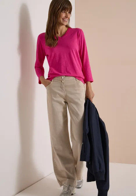 3/4-Arm Shirt in Unifarbe mit V-Neck pink flash Frau mit pinker Bluse, beigen weiten Hosen und weißen Schuhen hält eine dunkelblaue Jacke an hellem Hintergrund.