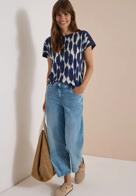 Frau in gemustertem T-Shirt, weiter Jeans, Wedges und mit geflochtener Tasche.