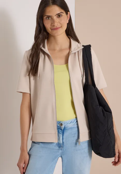 Frau in beiger Kurzarmjacke, gelbem Top, blauen Jeans und schwarzer Umhängetasche.