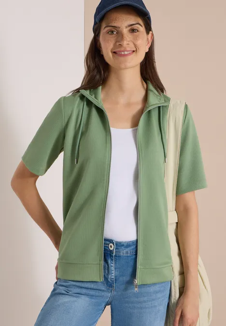 Frau in grüner Kurzarm-Hoodie-Jacke, weißem Top, Jeans, blauer Mütze und Tragetasche.