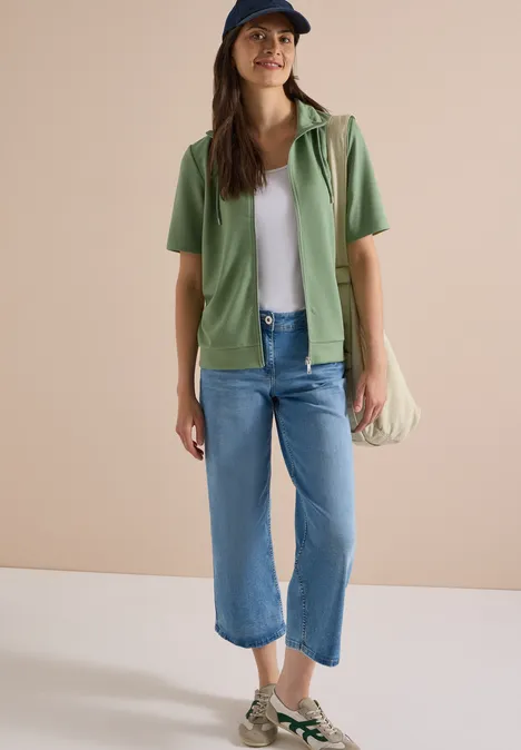 Frau in weißem Shirt, grüner Hemdjacke, blauen Jeans, Espadrilles und Schultertasche vor hellem Hintergrund.