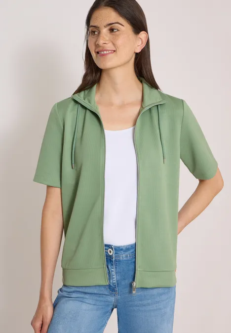 Damenmodell trägt grüne Kurzarm-Kapuzenjacke mit Reißverschluss, weißes Top und Jeans.