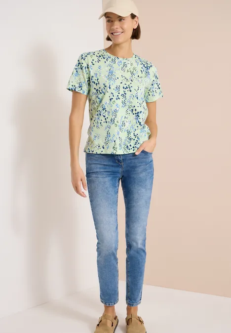 T-Shirt mit Rundhals und Print raw matcha lime Frau im Stehen trägt grün geblümtes T-Shirt, blaue Jeans und weiße Sneaker vor hellem Hintergrund.
