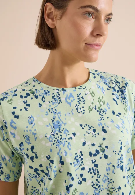 T-Shirt mit Rundhals und Print raw matcha lime Eine Frau in einem hellgrünen T-Shirt mit abstraktem blauem Fleckenmuster, schaut seitlich.