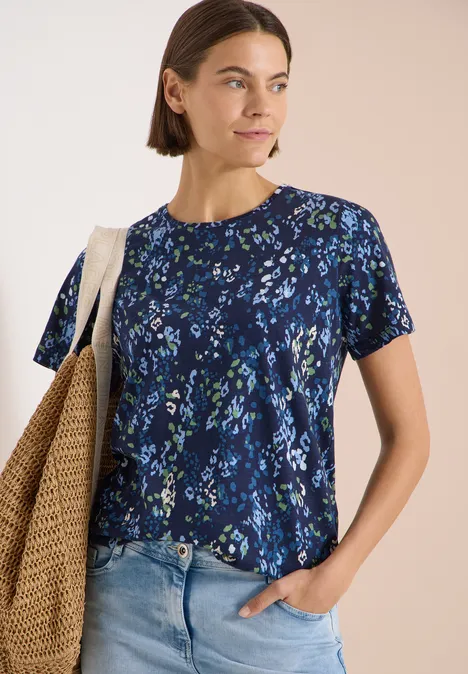 T-Shirt mit Rundhals und Print urban dark blue Frau trägt blaues T-Shirt mit Blumenmuster, halb in Jeans gesteckt, mit Strohtasche über der Schulter.
