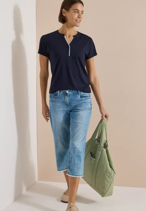 T-Shirt mit Split Neck in Unifarbe urban dark blue Frau in blauem T-Shirt mit Reißverschluss, weiten 3/4-Jeans, Beige-Slippern und grünem Steppbeutel.