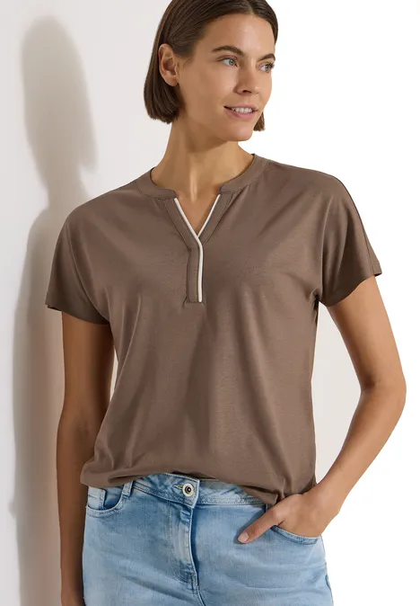 T-Shirt mit Split Neck in Unifarbe urban taupe Frau in braunem Kurzarmshirt mit weißem Stehkragen und Jeans lehnt an Wand und schaut seitlich.