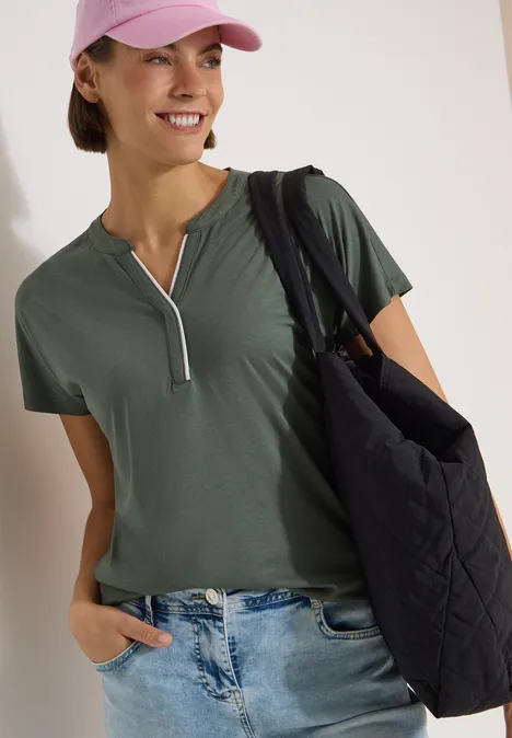 T-Shirt mit Split Neck in Unifarbe dried olive green Frau in grünem Kurzarmshirt mit Reißverschluss, Jeans, rosa Cap und großer schwarzer Stepp-Tasche.