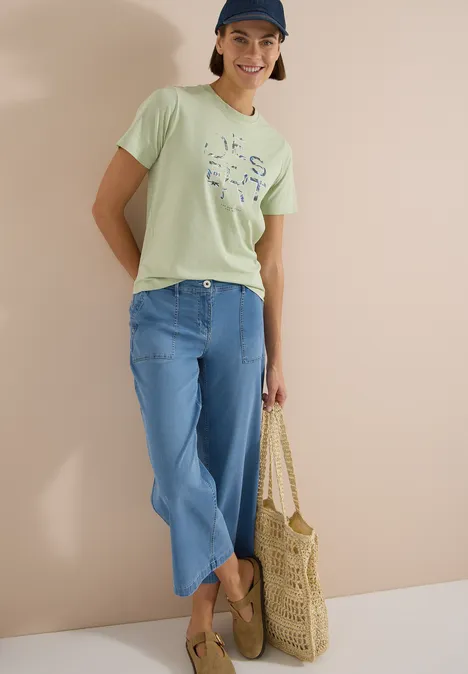 T-Shirt mit Rundhals und Frontprint raw matcha lime Person trägt hellgrünes Shirt, blaue Jeans, Basttasche und Slipper vor heller Wand.