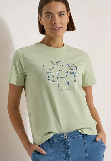 T-Shirt mit Rundhals und Frontprint raw matcha lime Frau in pastellgrünem T-Shirt mit abstraktem Print und blauen Jeans.