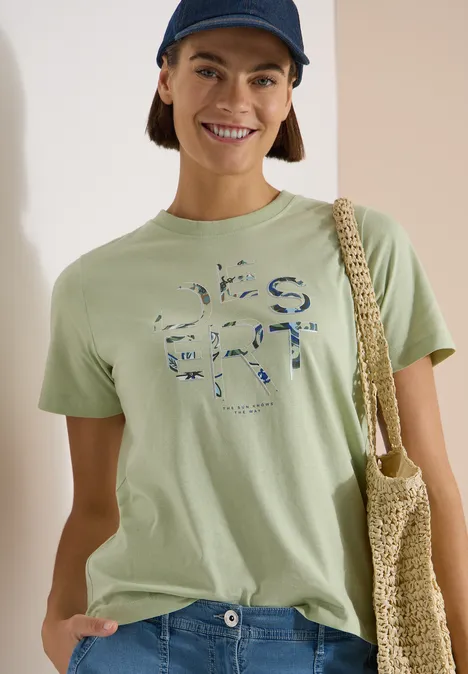 T-Shirt mit Rundhals und Frontprint raw matcha lime Eine Frau trägt ein hellgrünes T-Shirt mit Print, eine Netz-Tasche über der Schulter und eine dunkle Cap.