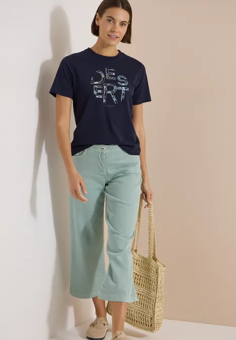 T-Shirt mit Rundhals und Frontprint urban dark blue Frau steht vor neutralem Hintergrund, trägt dunkelblaues T-Shirt mit Schriftzug, mintfarbene Culotte und Basttasche.