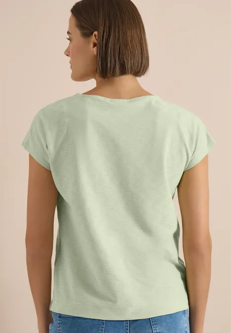 Personne vue de dos portant un T-shirt vert clair devant un fond neutre, cheveux au carré.