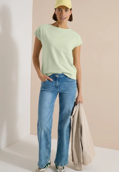 Femme en T-shirt vert pâle, jean droit bleu, tenant un manteau beige, sur fond épuré.