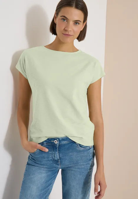 Femme portant un t-shirt vert clair aux manches retroussées et un jean bleu, main dans la poche.