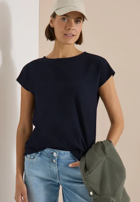 Femme porte T-shirt bleu marine et jean, tient une veste d'une main.