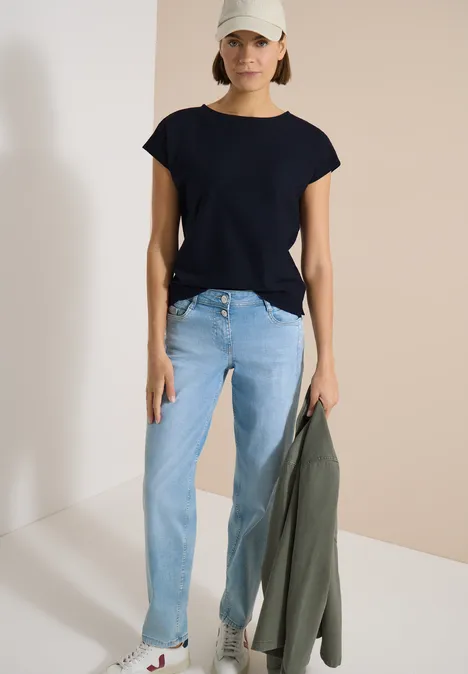 Femme en t-shirt noir et jean clair tenant une veste verte sur fond clair.