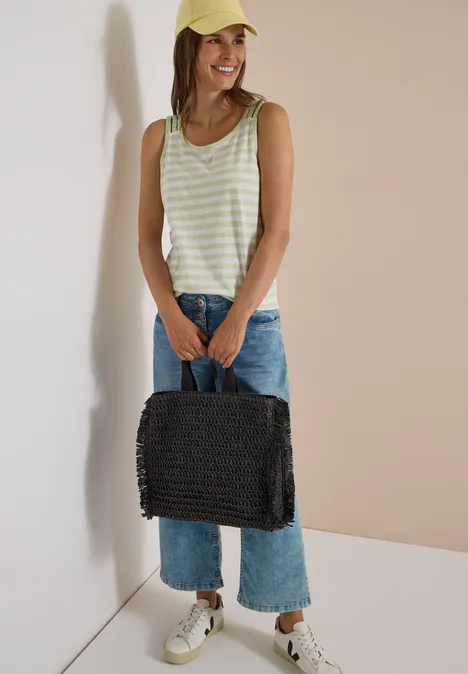 Junge Frau mit gelber Kappe, gestreiftem Top, Jeans und weißen Sneakern hält lächelnd eine schwarze Korbtasche.