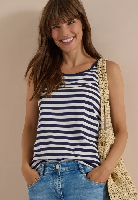 Lächelnde Frau in gestreiftem Top, Jeans und Strohtasche vor neutralem Hintergrund.