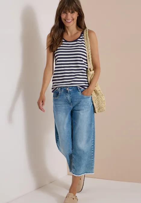 Junge Frau trägt gestreiftes Top, weite Jeans und Korbtasche, steht lächelnd an der Wand.
