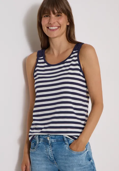 Frau in blau-weiß gestreiftem Tanktop und Jeans, lächelnd mit Händen in den Taschen.