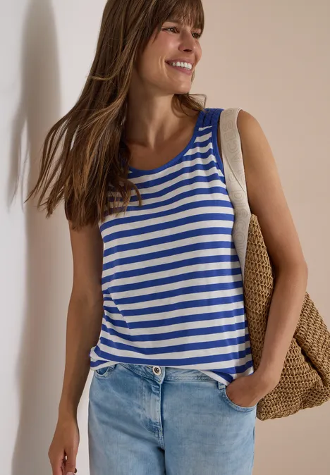 Femme adossée, top rayé bleu-blanc, jean et sac en osier, sourit détendue.