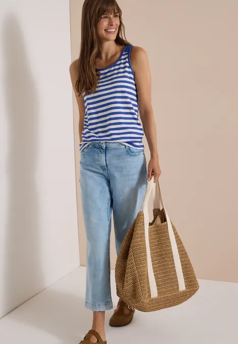 Femme souriante en top rayé et jeans bleus, tenant un grand sac en paille sur fond beige.