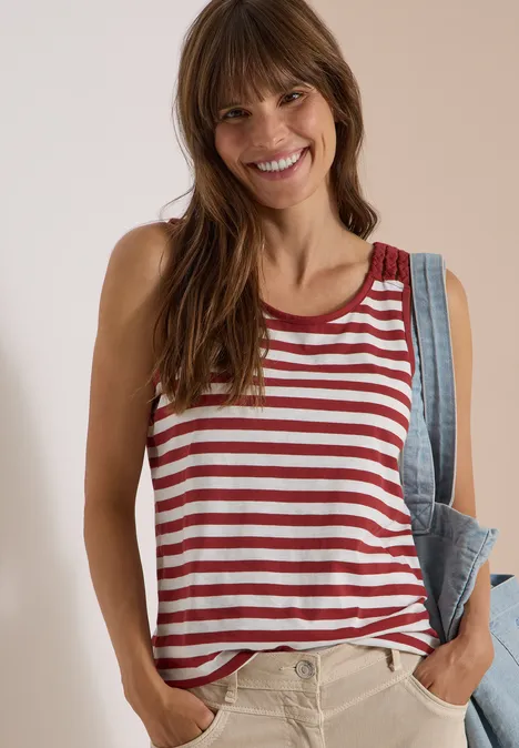 Femme souriante en débardeur rayé rouge et blanc, veste en jean posée sur l’épaule, mains dans les poches.