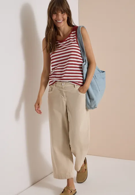 Une femme souriante porte un top rayé rouge et blanc, un pantalon large beige et un sac bleu sur fond clair.