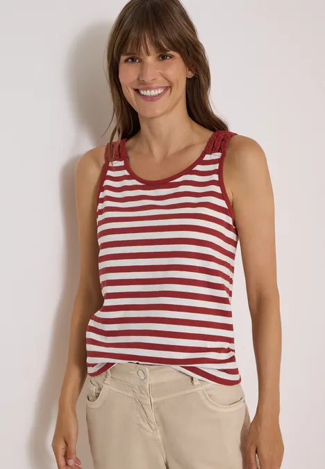 Femme souriante en débardeur rayé rouge et blanc, pantalon beige, devant un mur clair.