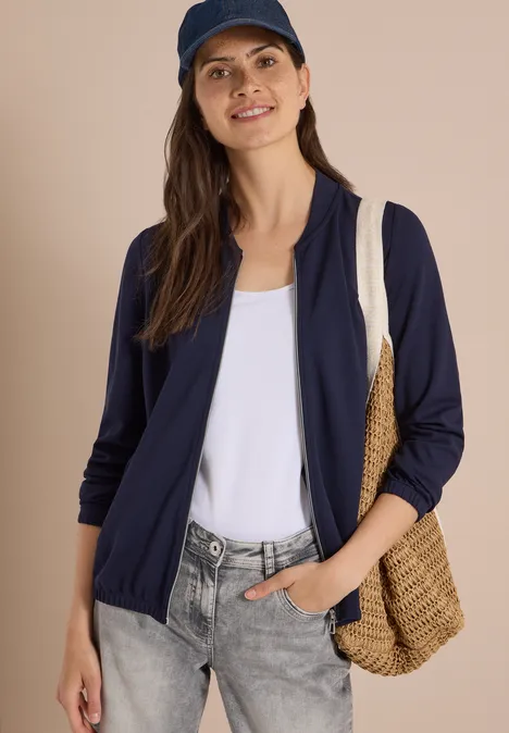 Frau in dunkelblauer Strickjacke, weißem Top, grauer Jeans, mit Strohtasche und Cap