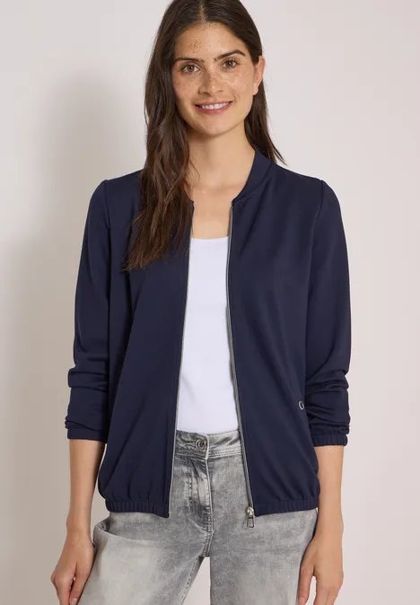 Frau trägt dunkelblaue Bomberjacke über weißem Top und grauen Jeans, elegante, lässige Optik.