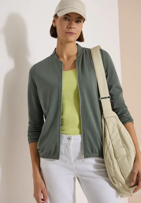 Langarm Jacke mit Rundhals und Zipper dried olive green Frau in grüner Jacke, limonengrünem Top, weißen Jeans und beiger gesteppter Tasche vor heller Wand.
