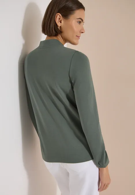 Langarm Jacke mit Rundhals und Zipper dried olive green Frau von hinten in grünem Langarmshirt und weißen Hosen, angelehnt an eine Wand.