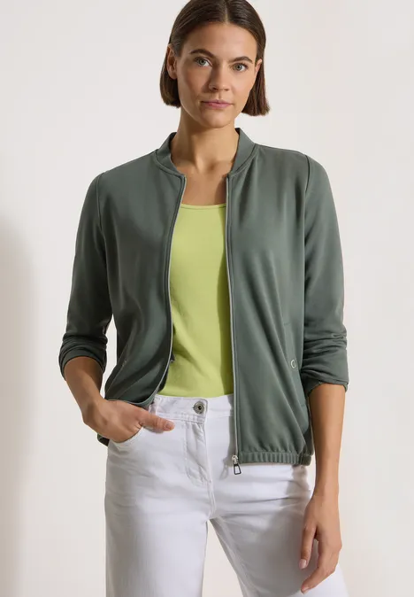 Langarm Jacke mit Rundhals und Zipper dried olive green Frau trägt khakigrüne Jacke über hellgrünem Top und weiße Hose, Hände in den Taschen.