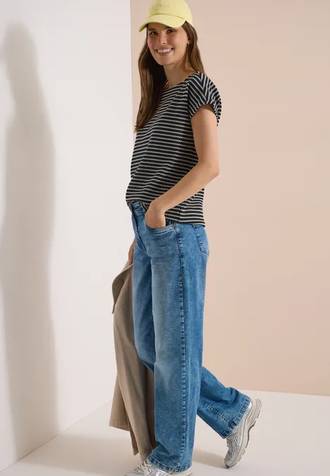 Een vrouw in een gestreept t-shirt, wijde jeans en sneakers staat nonchalant met een jas in haar hand.