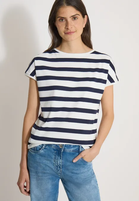 Model draagt wit-blauw gestreept T-shirt met lichte jeans, staat nonchalant met één hand in de zak.