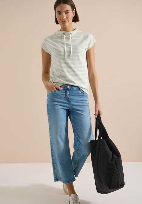 T-Shirt mit Funnel Neck und Streifen raw matcha lime Frau in hellgrauem Kapuzen-Shirt, weitem 7/8-Jeans und schwarzer Stepp-Tasche vor hellem Hintergrund.