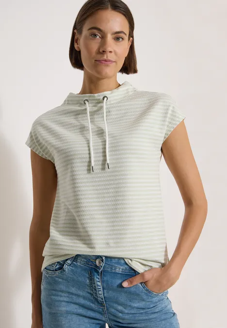T-Shirt mit Funnel Neck und Streifen raw matcha lime Frau in gestreiftem kurzärmeligen Kapuzenshirt mit Kordelzug und Jeans vor neutralem Hintergrund.