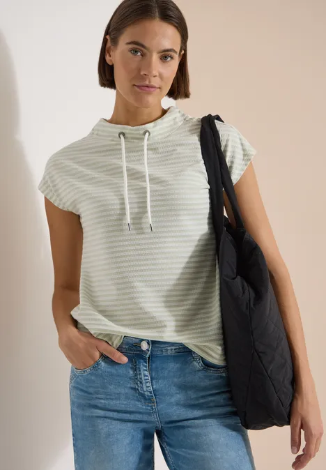 T-Shirt mit Funnel Neck und Streifen raw matcha lime Frau mit gestreiftem Kurzarm-Hoodie, Jeans und schwarzer gesteppter Umhängetasche.