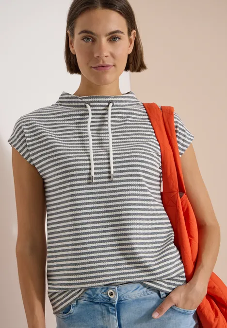 T-shirt à col cheminée et rayures urban dark blue Person trägt gestreiftes Ärmelloses Kapuzenshirt, Jeans, hält eine orangene Tasche, vor beigem Hintergrund.